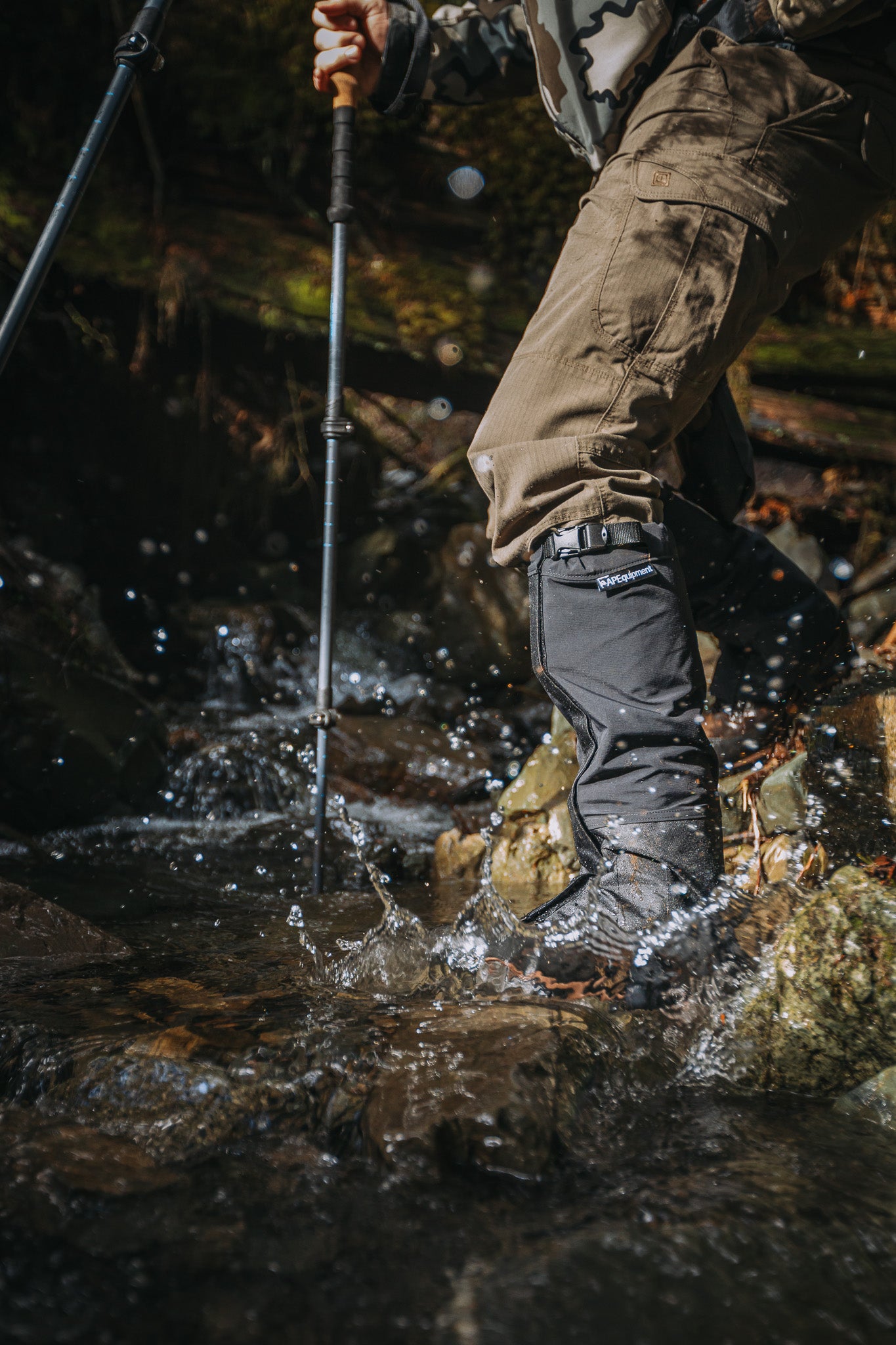 Person hiking through a stream wearing brown pants and black boots with a walking stick.