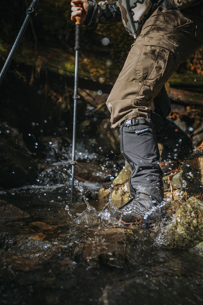 Person hiking through a stream wearing brown pants and black boots with a walking stick.